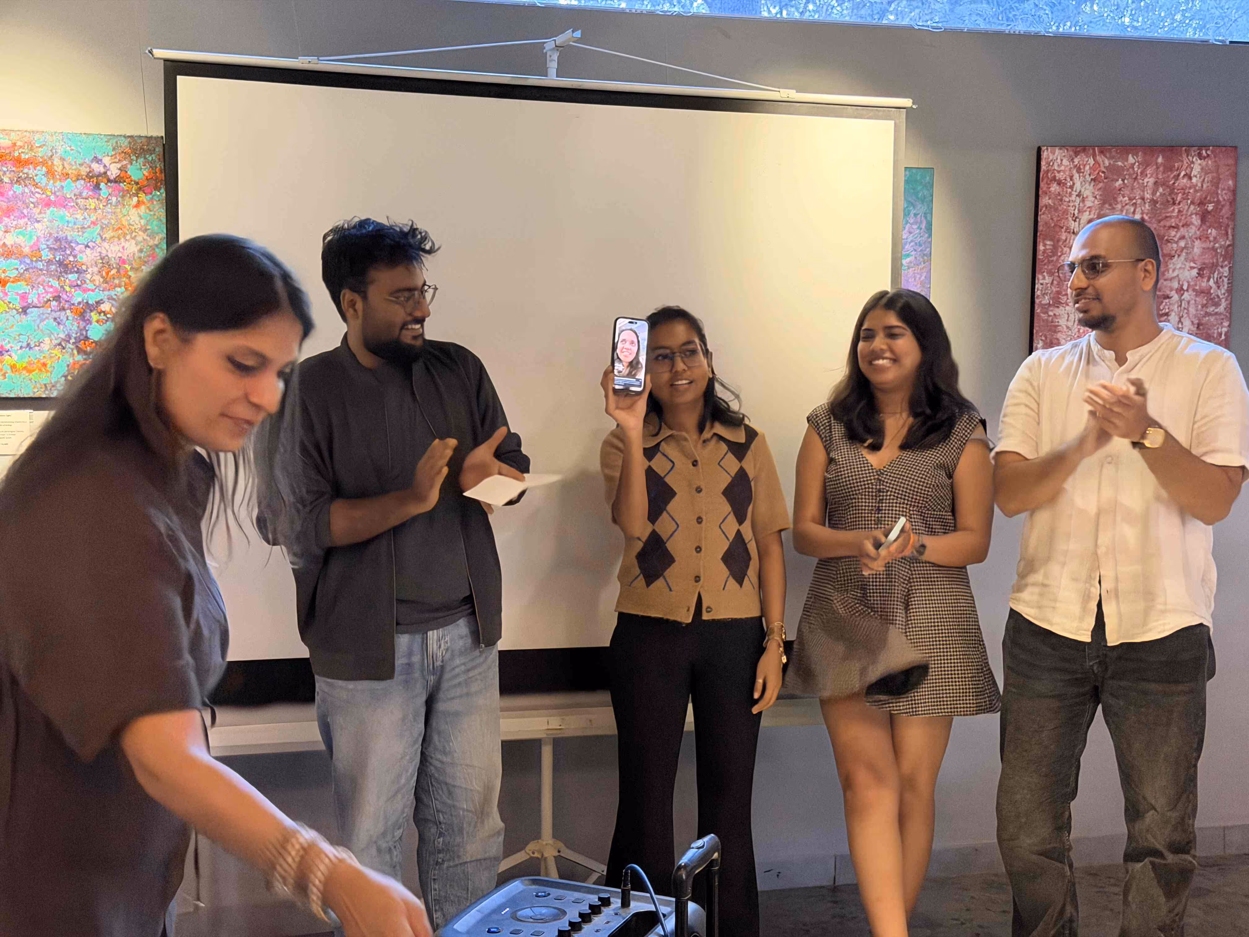 Five people stand in front of a projector screen; one holds up a smartphone showing a sixth person on a video call, while others clap and smile.