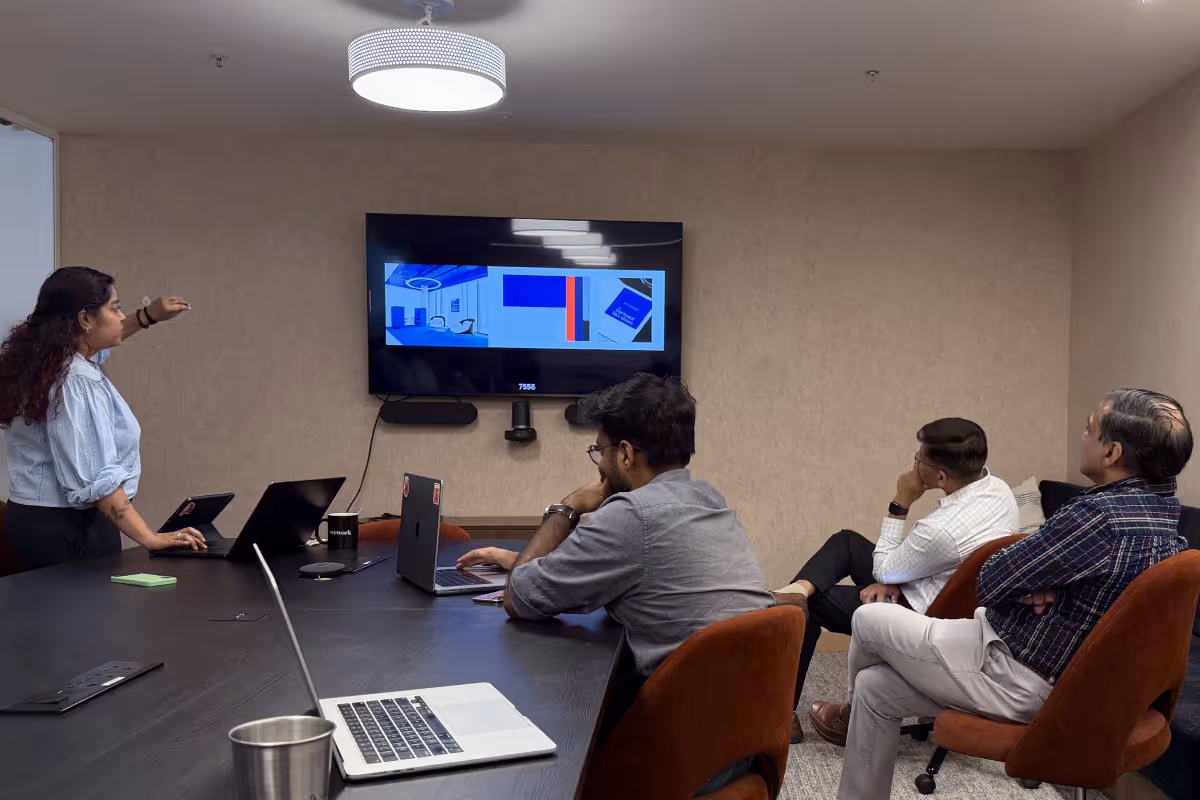 A woman standing and presenting on a screen to three seated men in a small conference room with laptops.