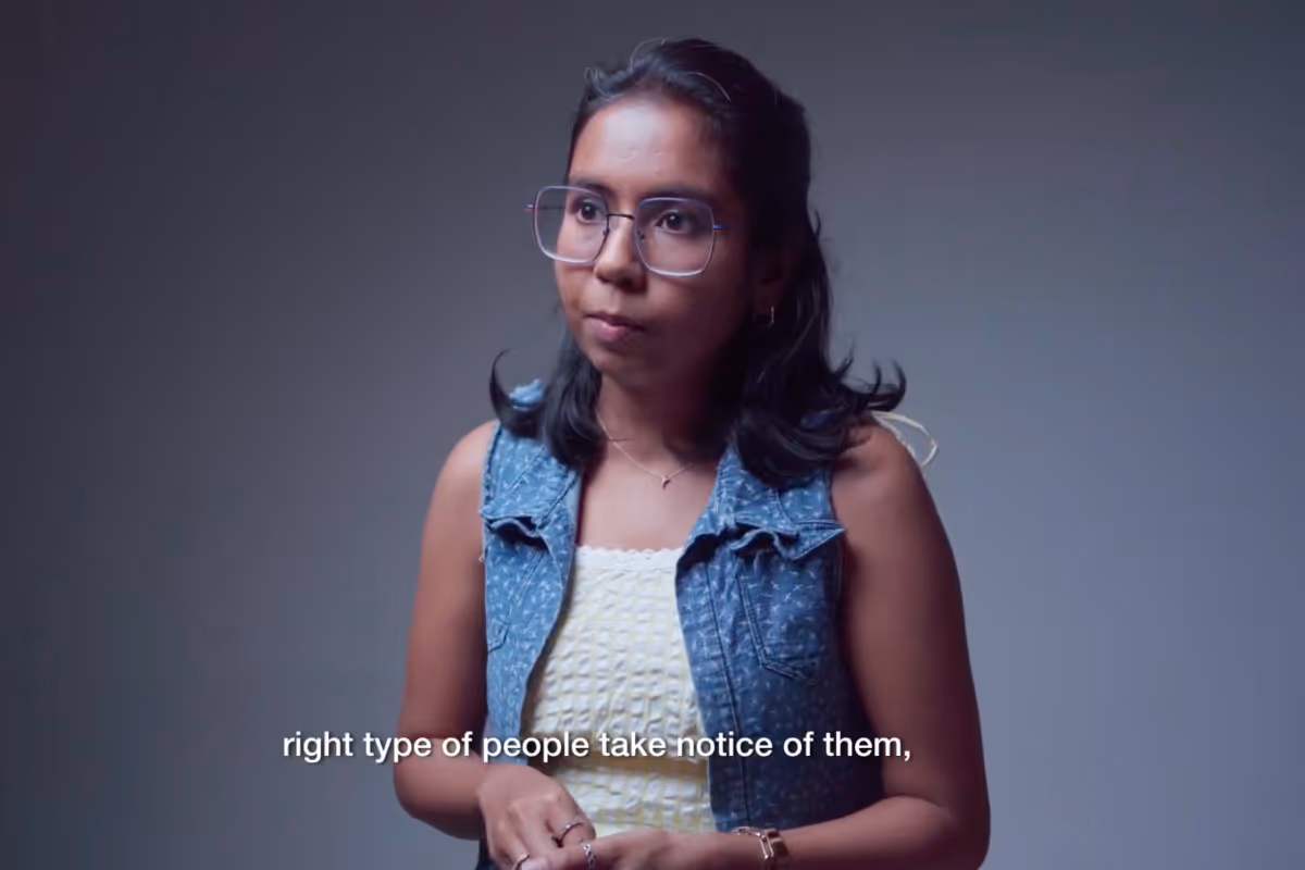 Woman with glasses and a blue sleeveless denim vest over a yellow top speaking with subtitle text 'right type of people take notice of them,' on a gray background.