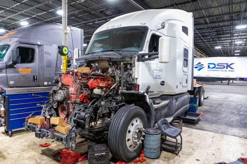 A brightly lit truck repair bay with a blue Kenworth semi-truck. Tools, bins, and supplies are organized between them. A mechanic works in the background. “Truck Bay 7” signage hangs above the well-maintained workspace.