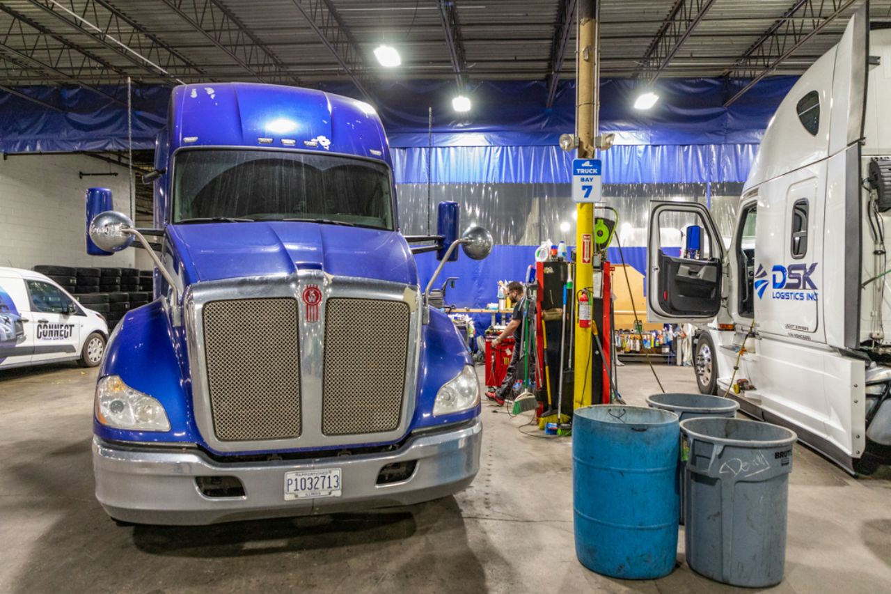 A brightly lit truck repair bay with a blue Kenworth semi-truck. Tools, bins, and supplies are organized between them. A mechanic works in the background. “Truck Bay 7” signage hangs above the well-maintained workspace.