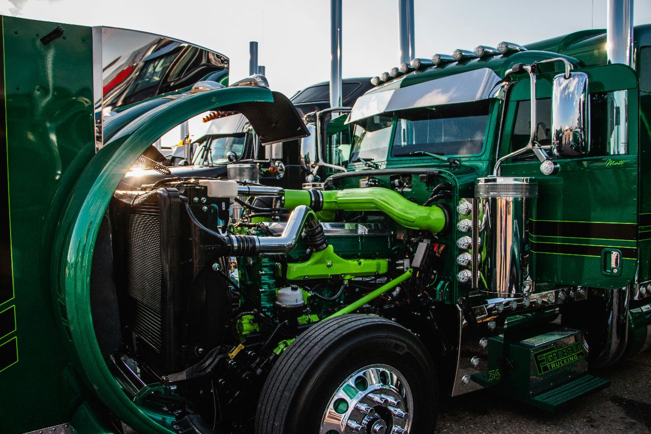 Green semi truck with hood open, exposing detailed diesel engine and chrome components at truck show in daylight.