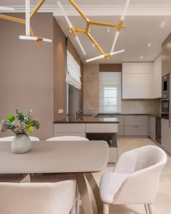 Modern kitchen with beige and gray cabinetry, a central island, minimalist light fixtures, and a dining table with upholstered chairs and a floral centerpiece.