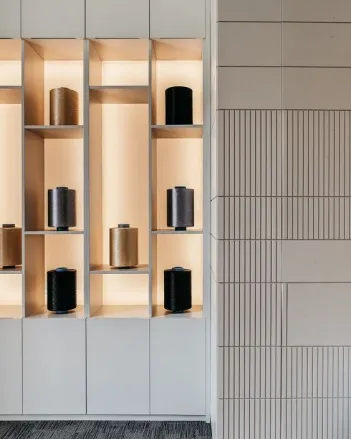 Modern shelving unit with illuminated cubic compartments holding various large spools of thread in black, brown, and beige colors, next to a textured wall with vertical and horizontal lines.