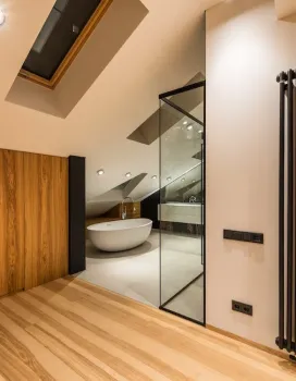 Modern bathroom with a freestanding white bathtub under a slanted ceiling with a skylight, visible through a glass partition, next to a wooden floor and wall panel.