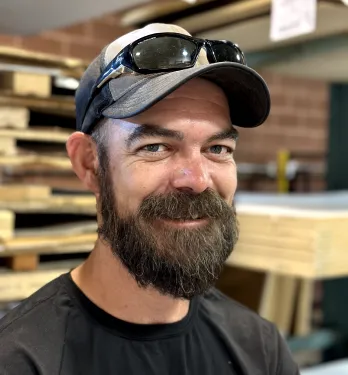 Bearded man wearing a cap with sunglasses on top, smiling in a workshop with stacked wooden planks in the background.