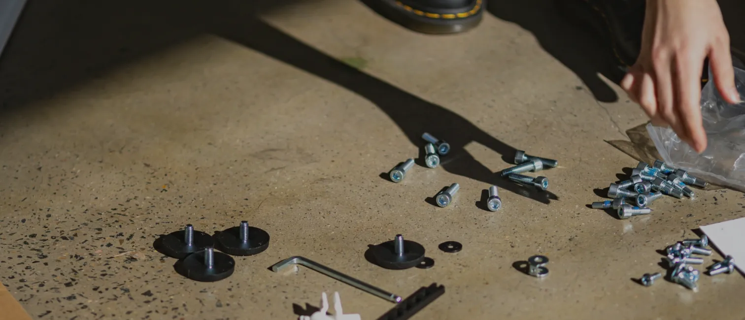 Various screws, bolts, and an Allen wrench scattered on a concrete floor with a hand reaching toward a plastic bag.