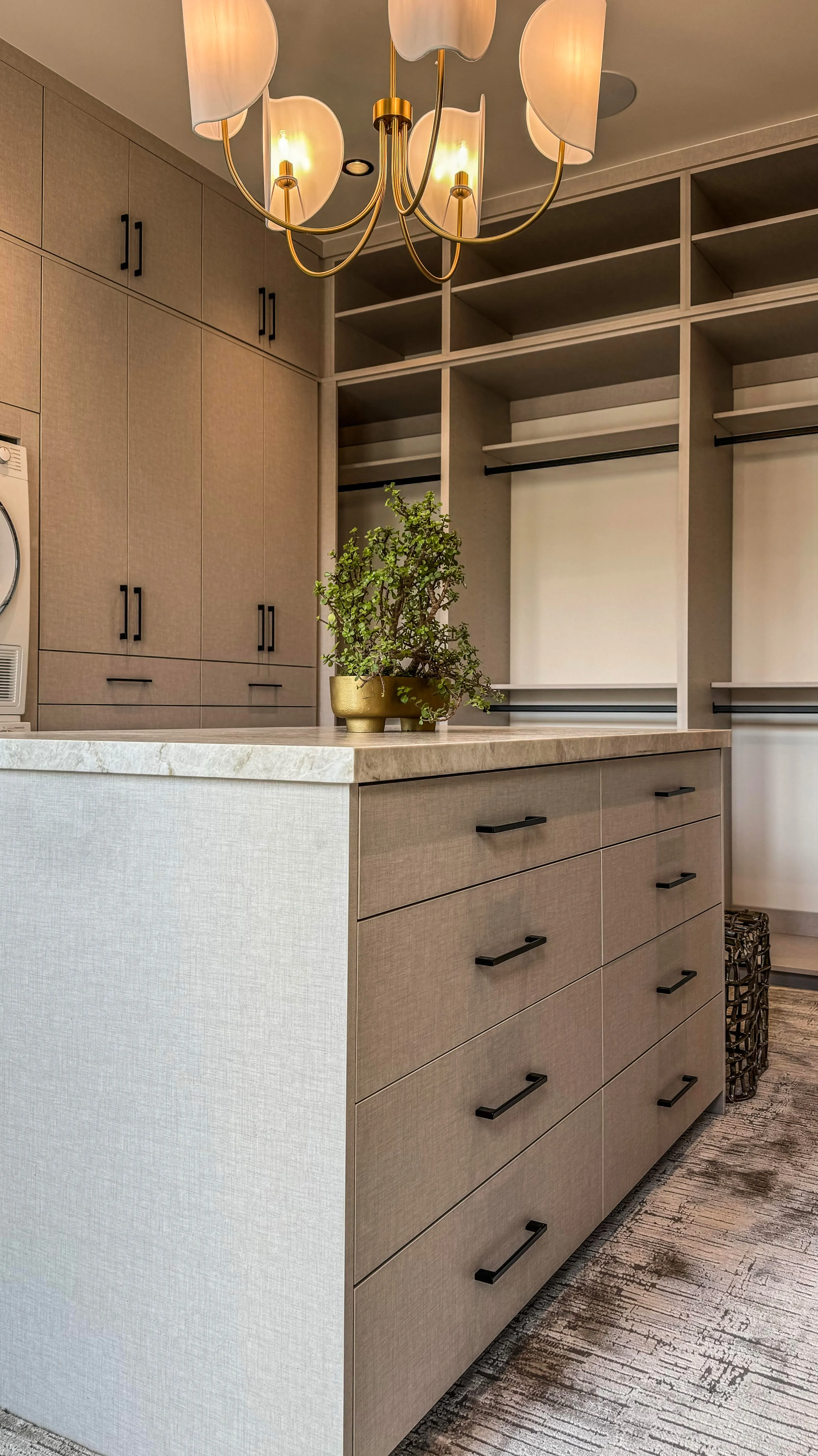 Modern walk-in closet with beige cabinetry, island drawers, open shelves, and a gold chandelier above.