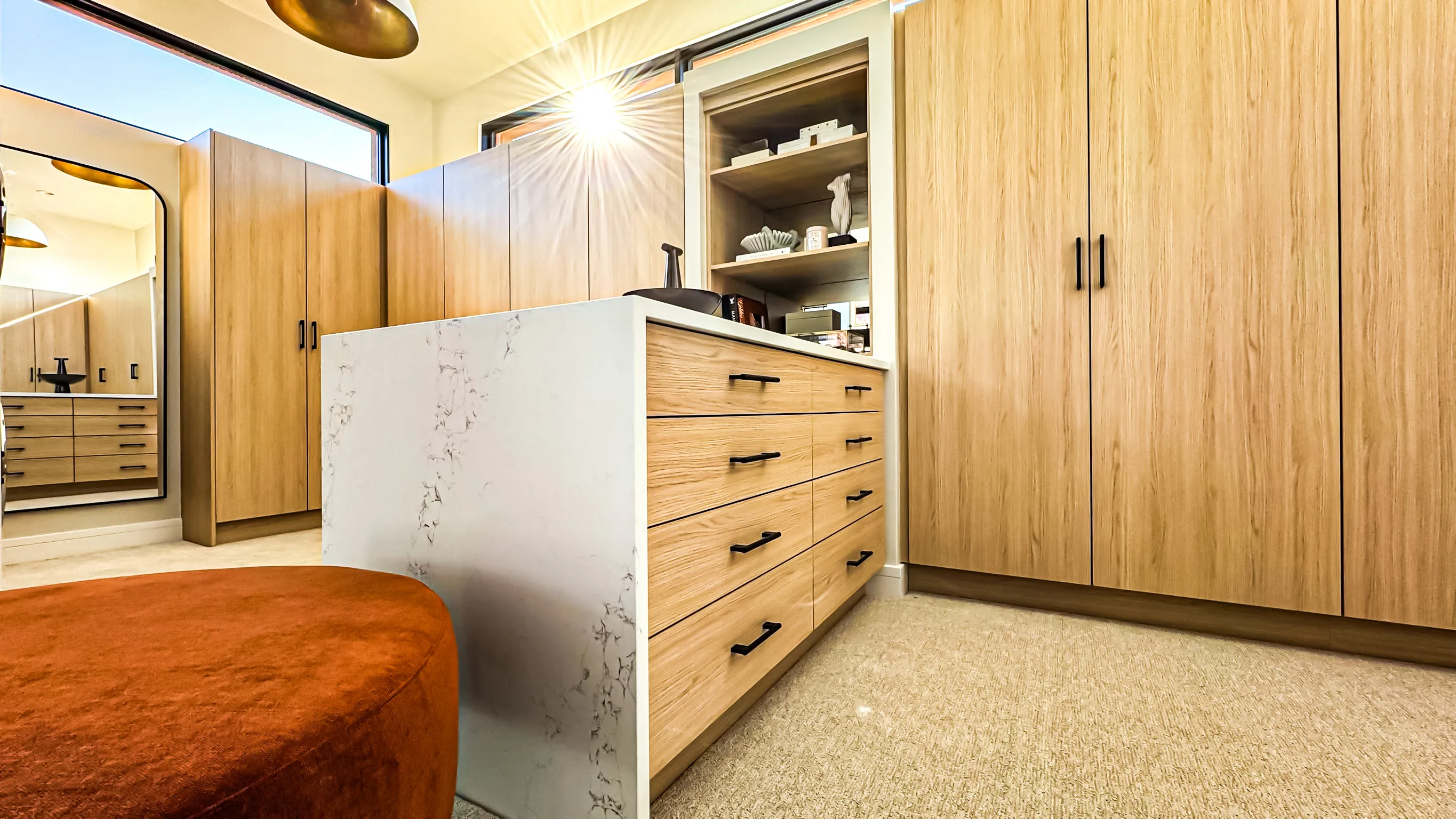 Modern walk-in closet with light wood cabinetry, marble-topped island drawers, beige carpet, and a large mirror reflecting the room.