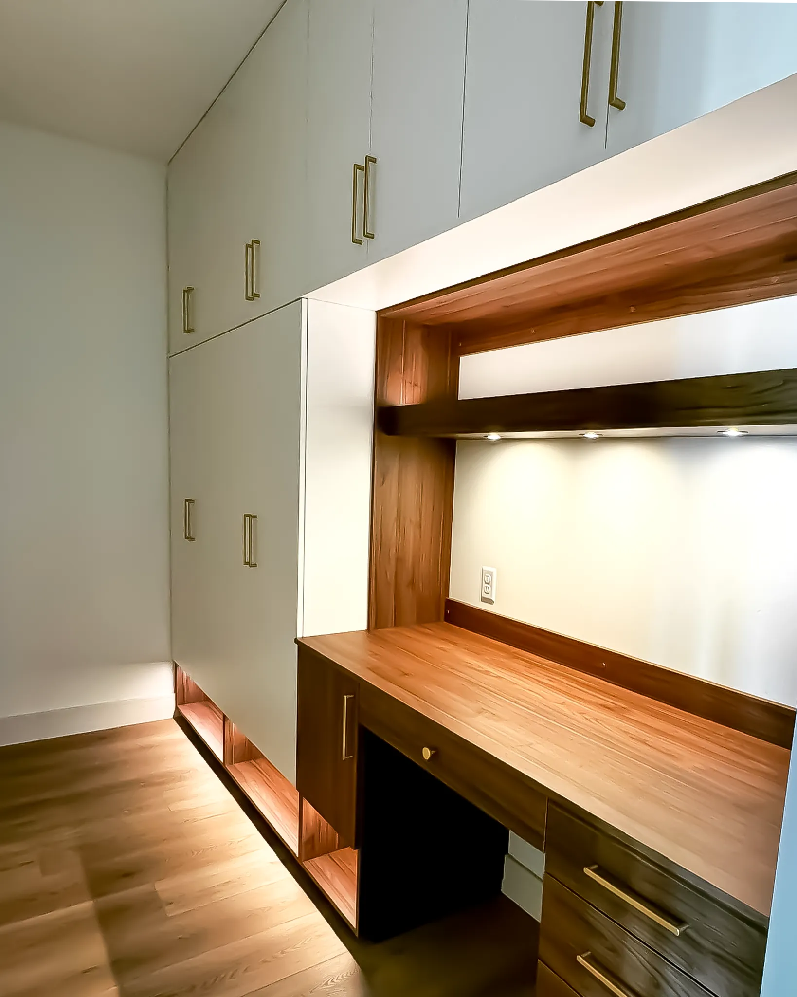Modern built-in wooden desk with overhead lighting and white cabinets above and beside it on wooden floor.
