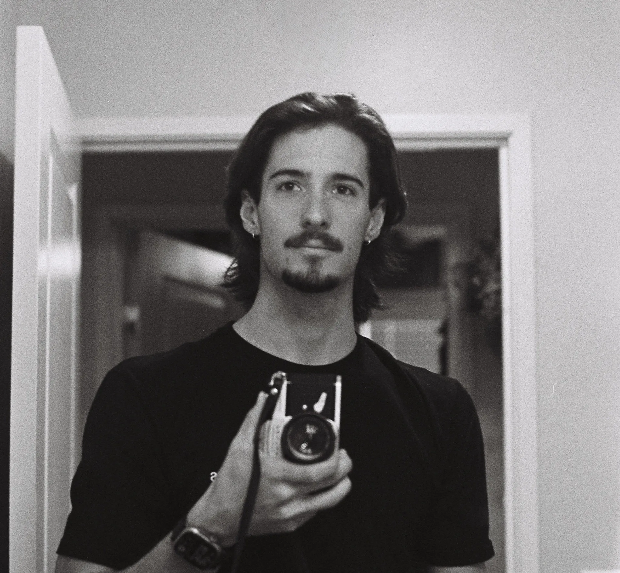 Black-and-white selfie of a young man with medium-length hair and a goatee holding a camera in front of a mirror.
