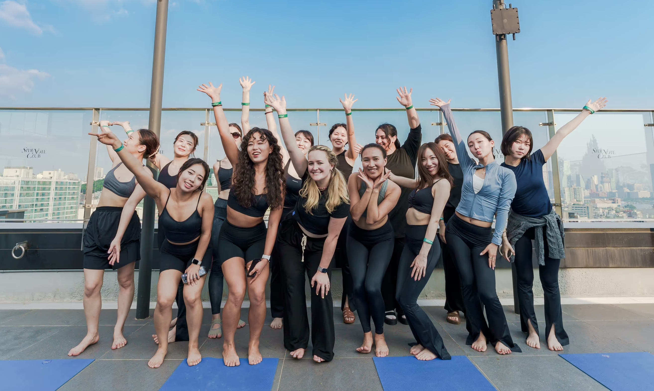 Group of diverse women posing cheerfully on yoga mats outdoors with city skyline in the background.