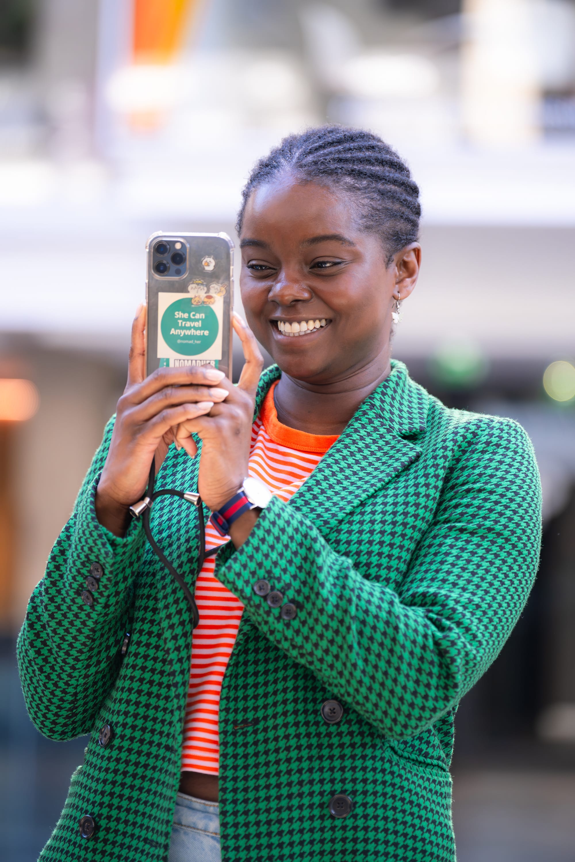 Smiling woman with braided hair in green houndstooth blazer and orange striped shirt holding phone with a sticker reading 'She Can Travel Anywhere'.