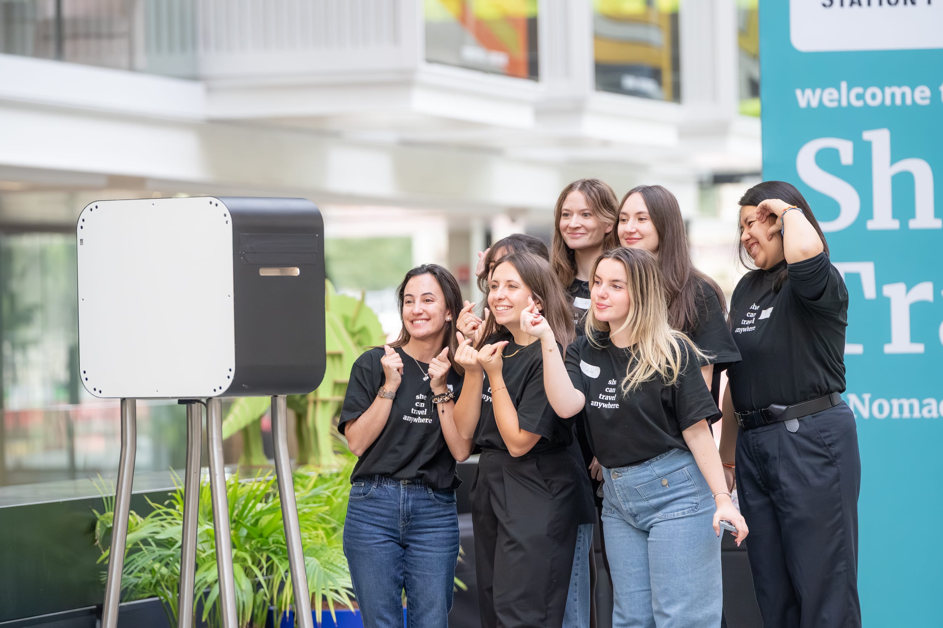 Group of six young women wearing matching black shirts posing together and smiling in front of a photo booth indoors.