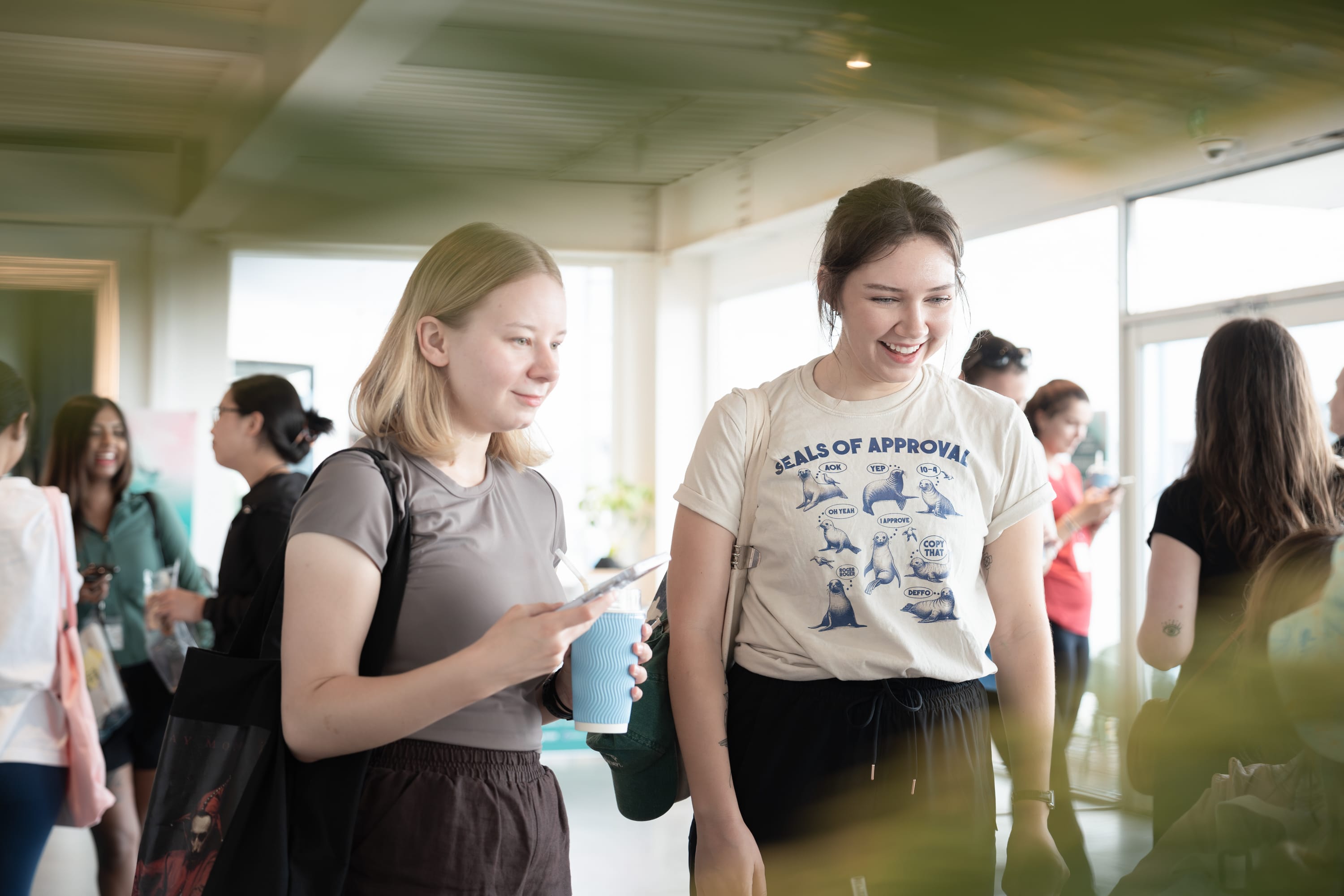 Two young women smiling and holding drinks while talking in a bright room with others in the background.