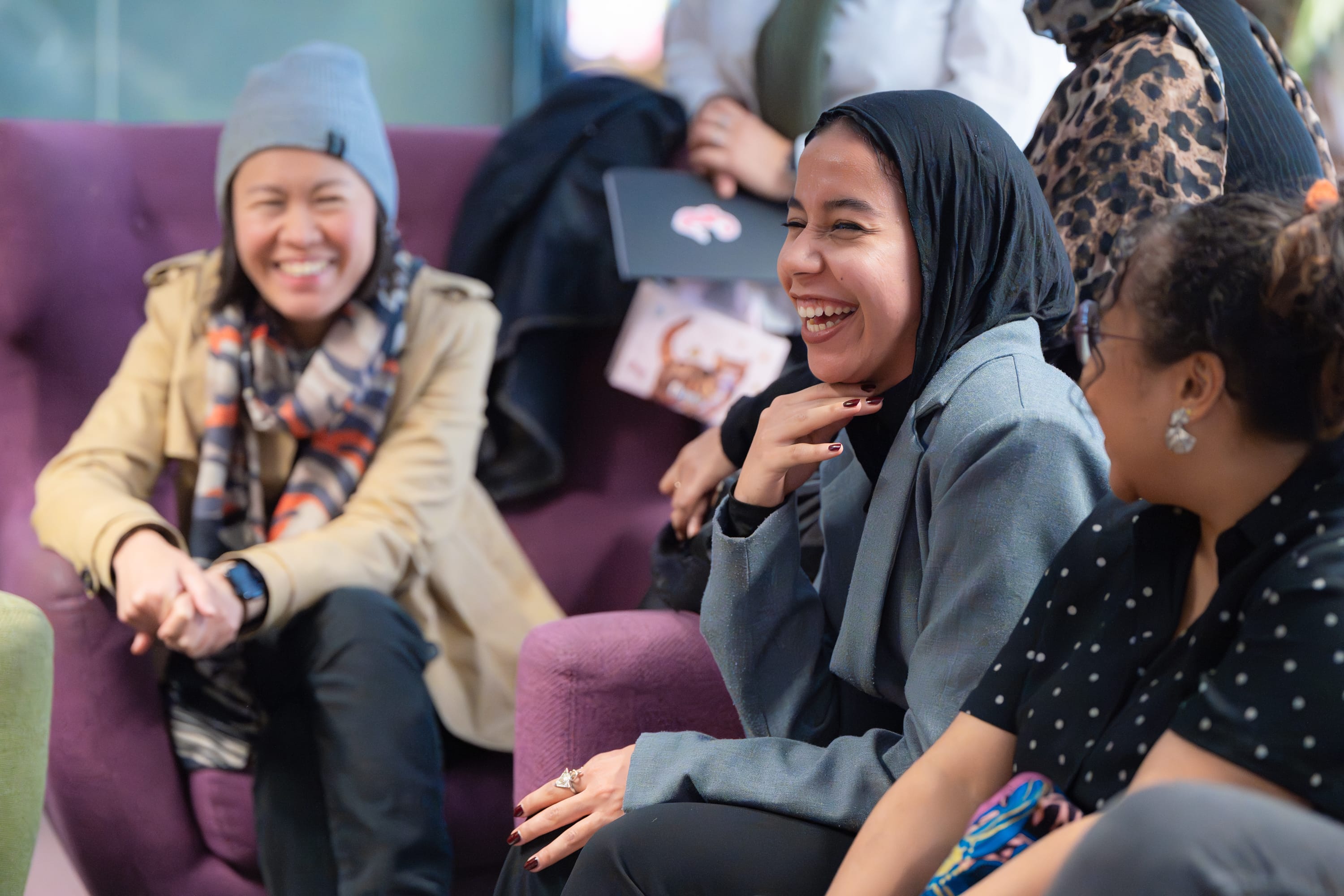 Three women sitting on a purple couch, smiling and laughing together in a casual setting.