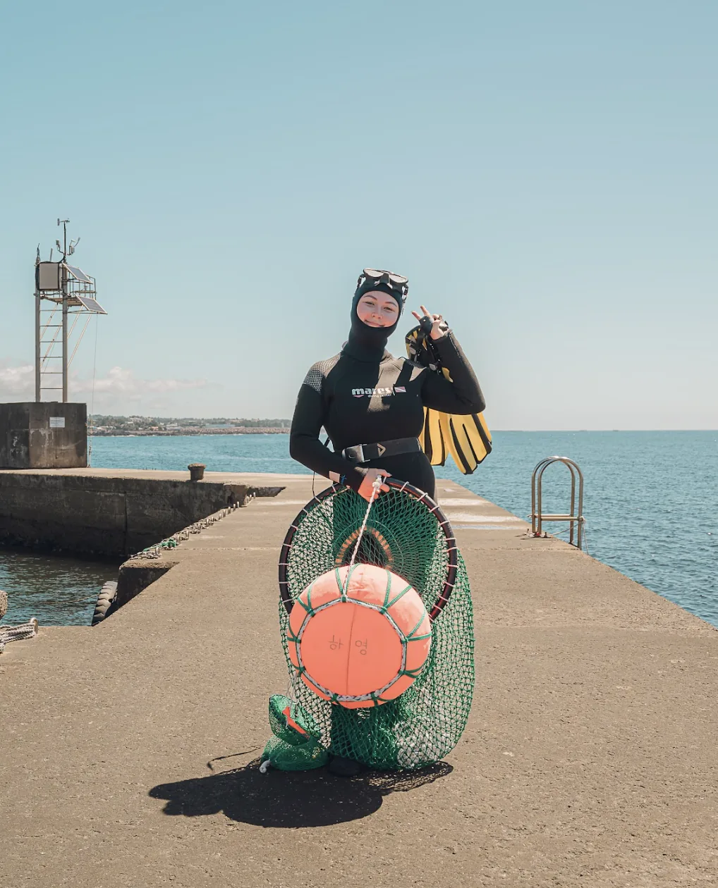 A woman host in a wetsuit holding a diving net on a harbour pier
