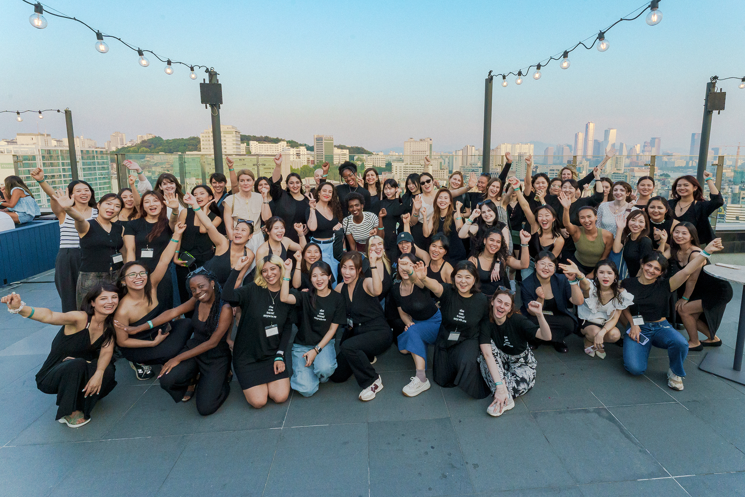 Large diverse group of women smiling, cheering, and raising fists together on a rooftop with city buildings and string lights in the background during daylight.