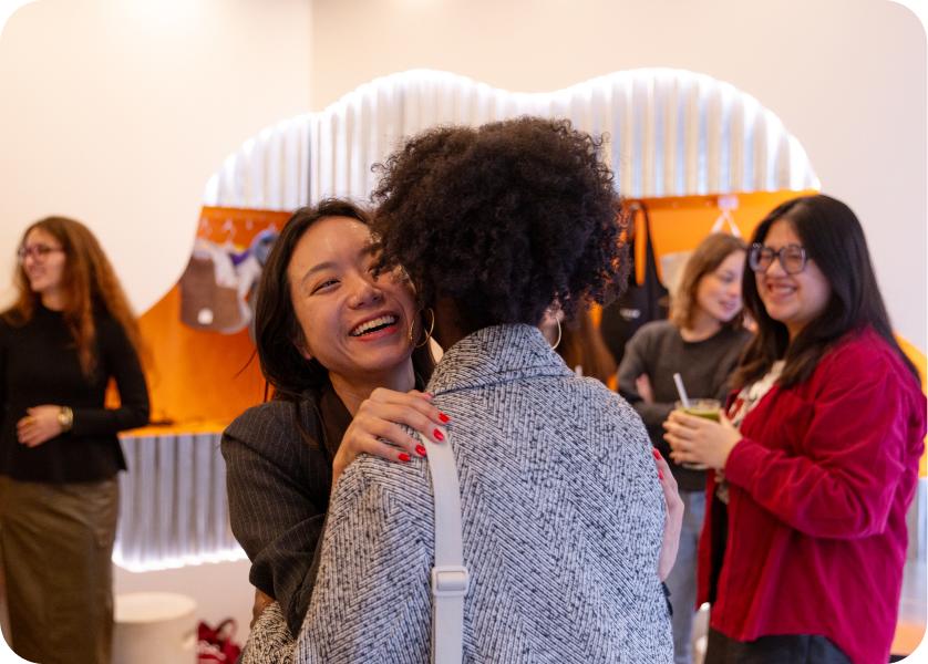 Two women warmly hug and smile in a social setting while three other women chat and smile in the background at a casual indoor gathering.