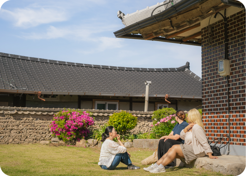 Three women sitting outside a traditional-style house with a tiled roof and brick wall, surrounded by green grass, blooming pink and purple flowers, and bushes under a blue sky.