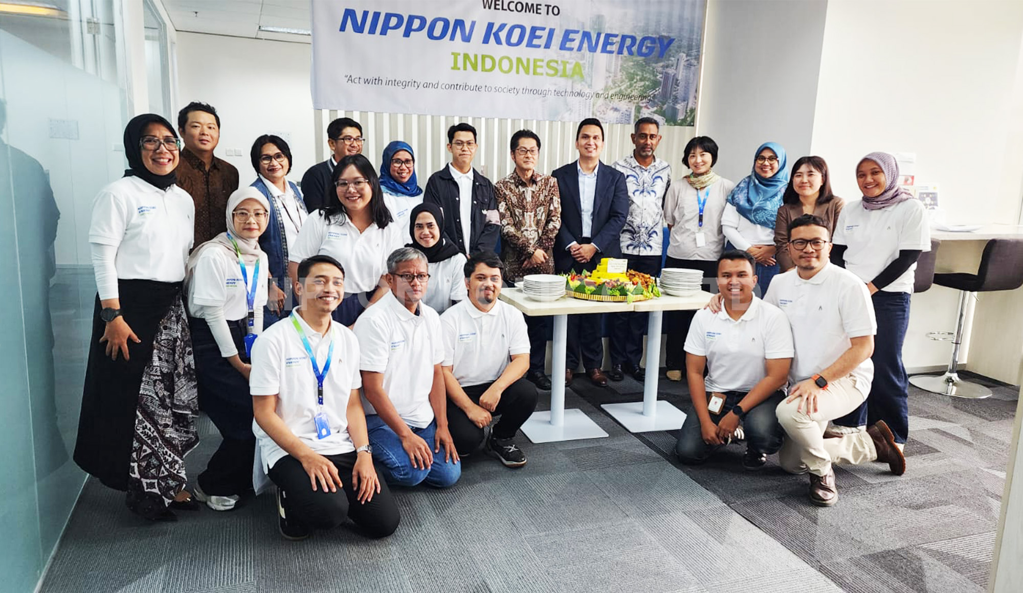 Group of employees smiling and posing in an office with a table of food and a banner that reads 'Welcome to Nippon Koei Energy Indonesia'.