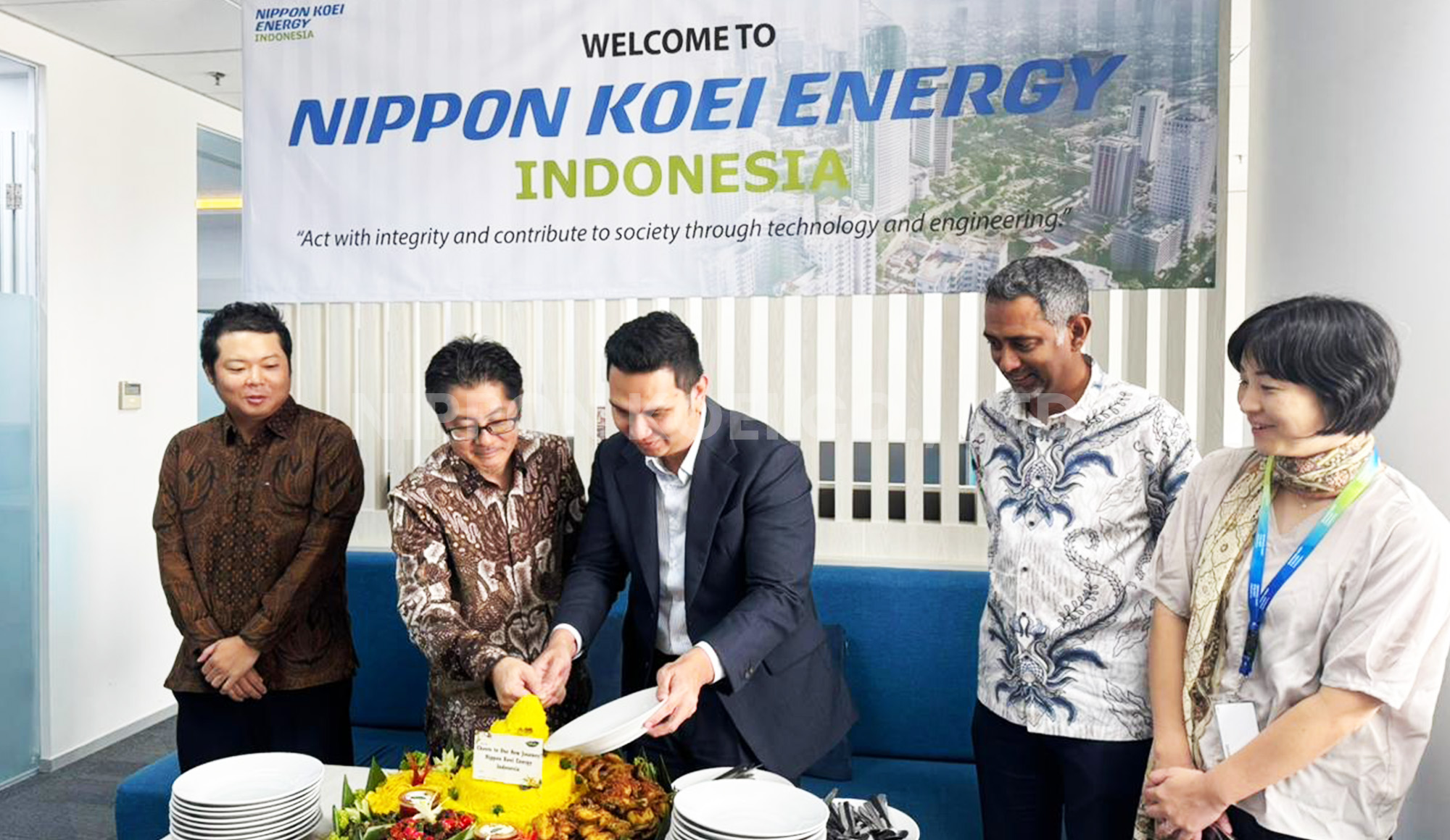 Five people standing around a table with a festive yellow rice cone, with two wearing batik shirts and a banner reading Nippon Koei Energy Indonesia in the background.