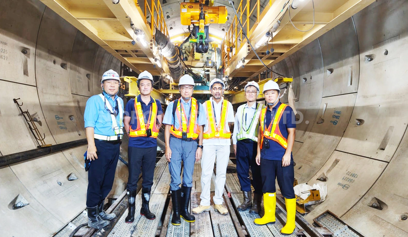 Six workers in safety gear standing inside a tunnel construction site with machinery overhead.