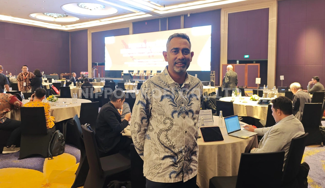 Man in patterned shirt stands in a conference room with round tables and seated attendees using laptops.