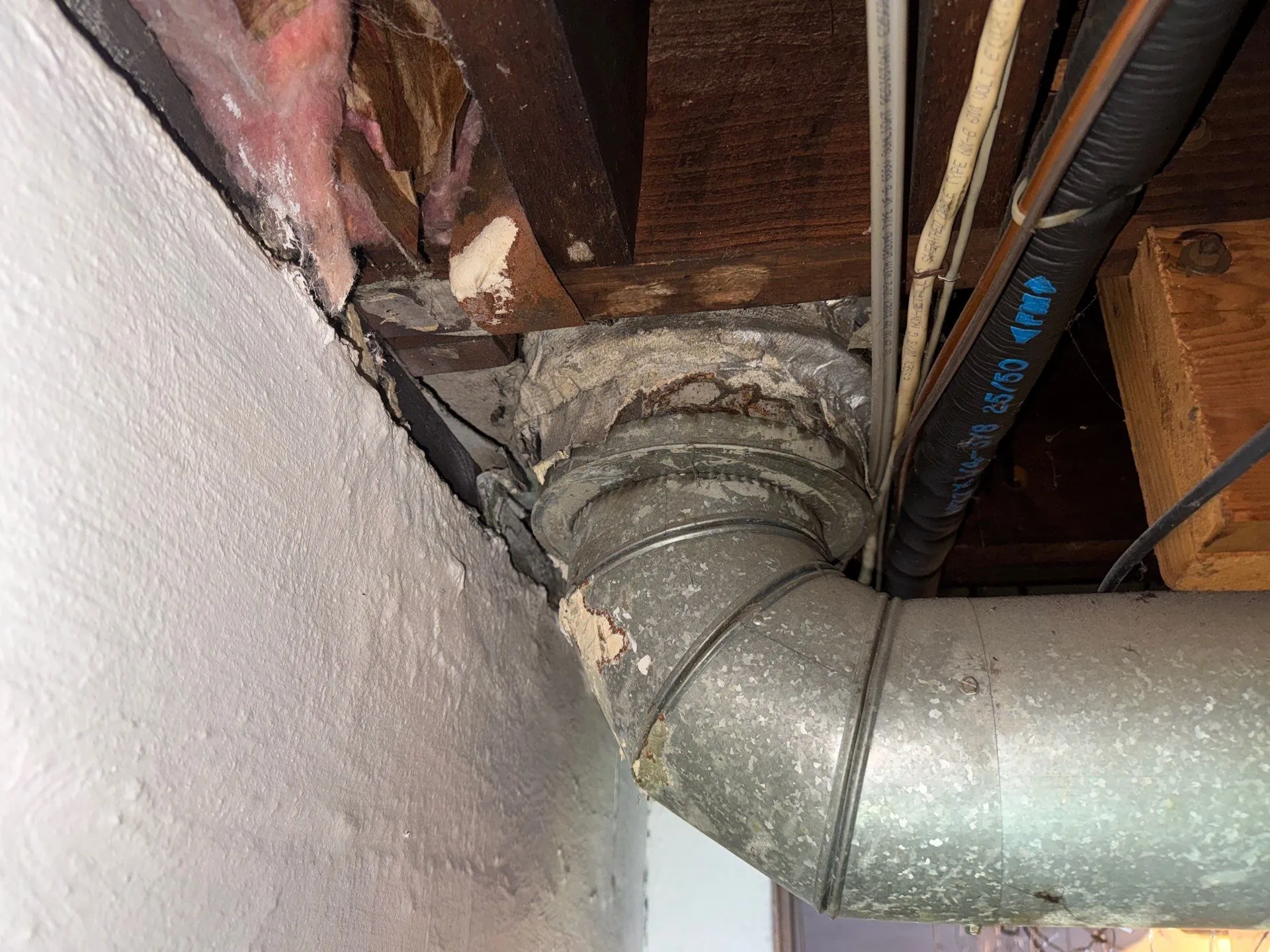 Metal duct pipe with worn insulation and pink fiberglass insulation near wooden beams and white wall corner.