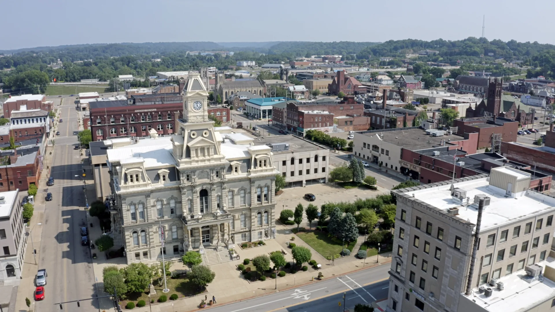 Aerial view of a small downtown area featuring a prominent historic courthouse with a clock tower, surrounded by streets, parked cars, trees, and brick buildings, with green hills in the background under a clear sky.