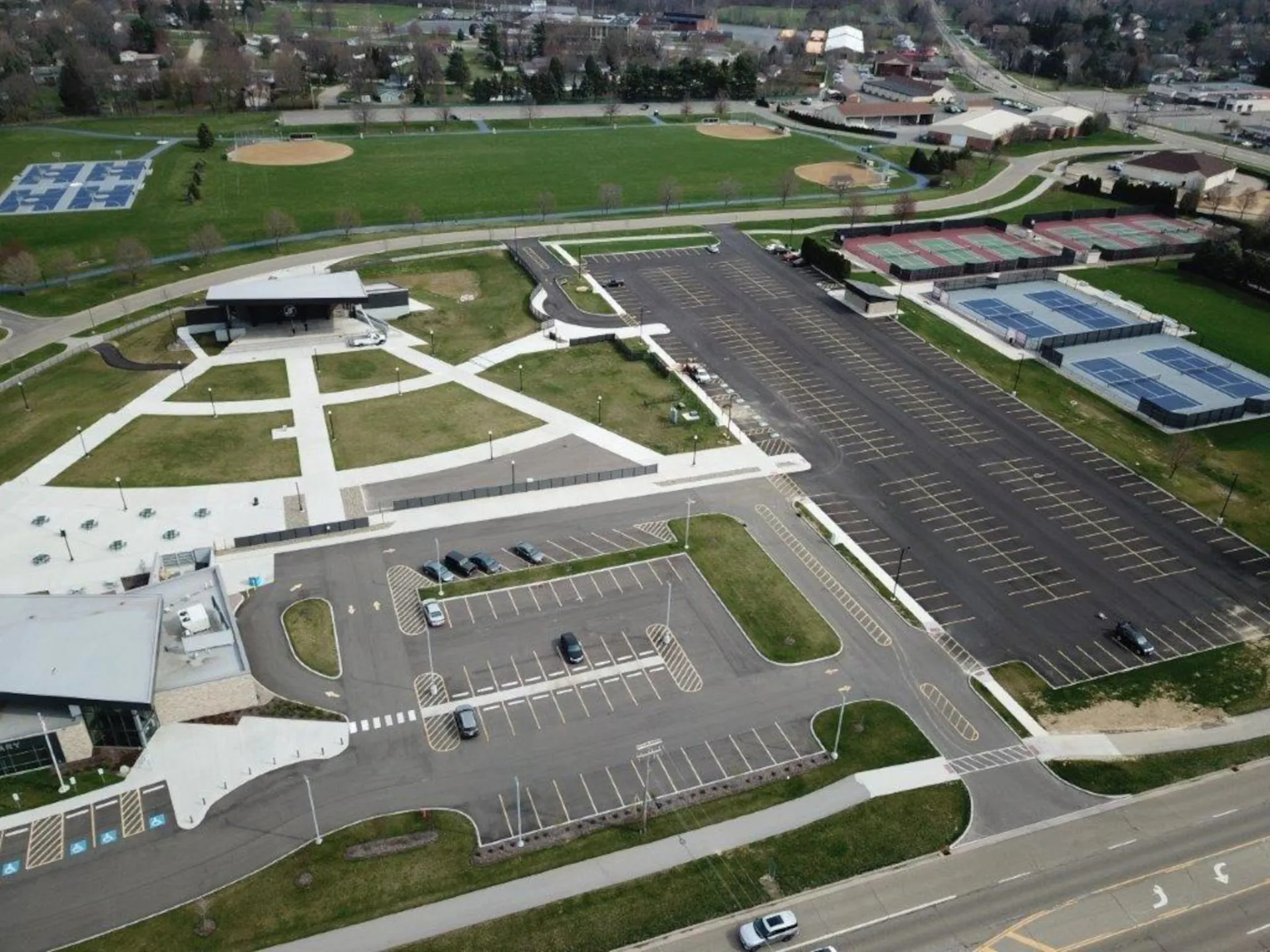 Aerial view Jackson North Park in Northeast Ohio, showing parking lots, tennis courts with red and blue surfaces, baseball fields, a building with walkways, and surrounding roads and trees.