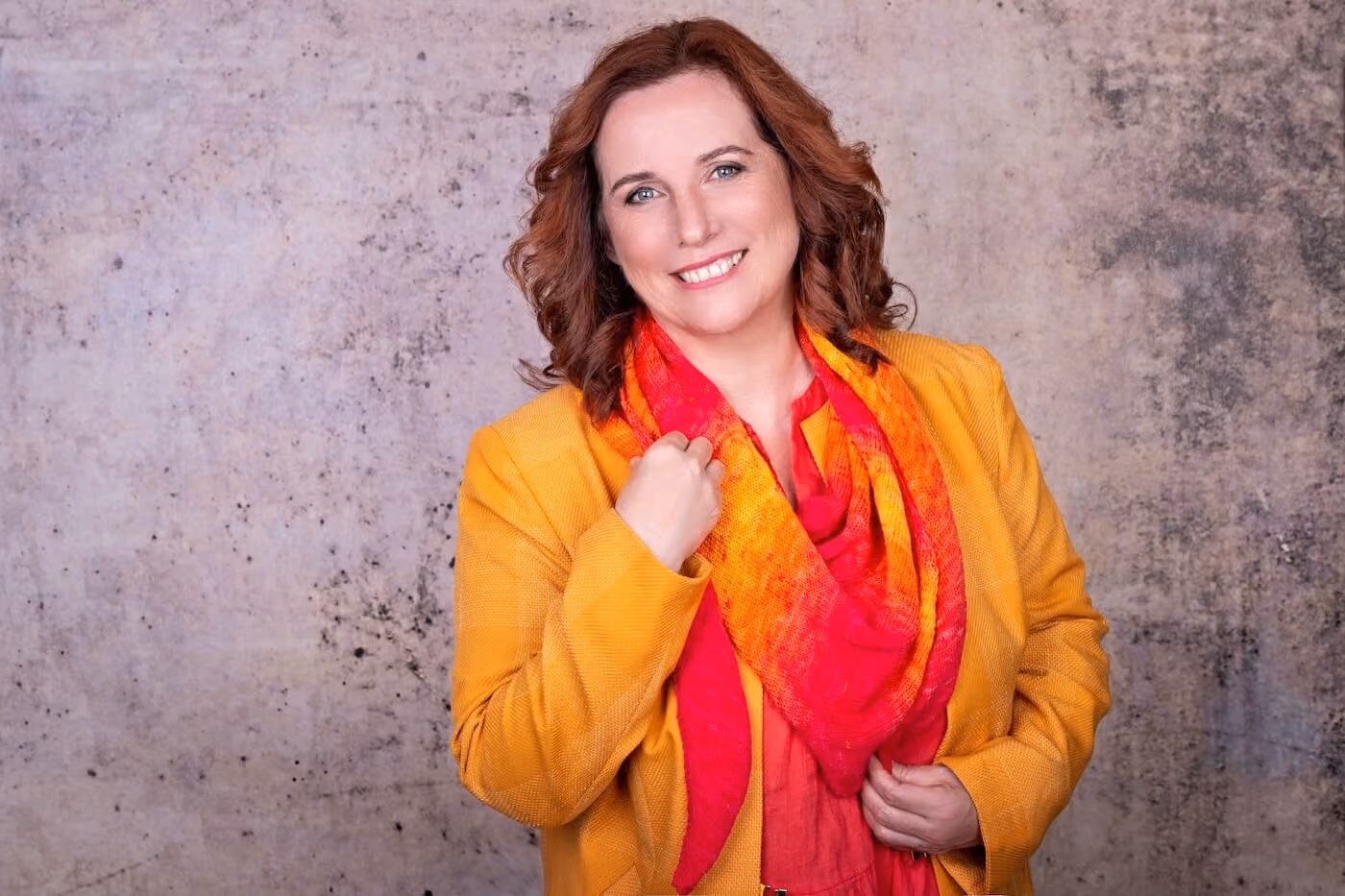 Smiling woman with red hair wearing a yellow jacket and red-orange scarf against a textured wall.