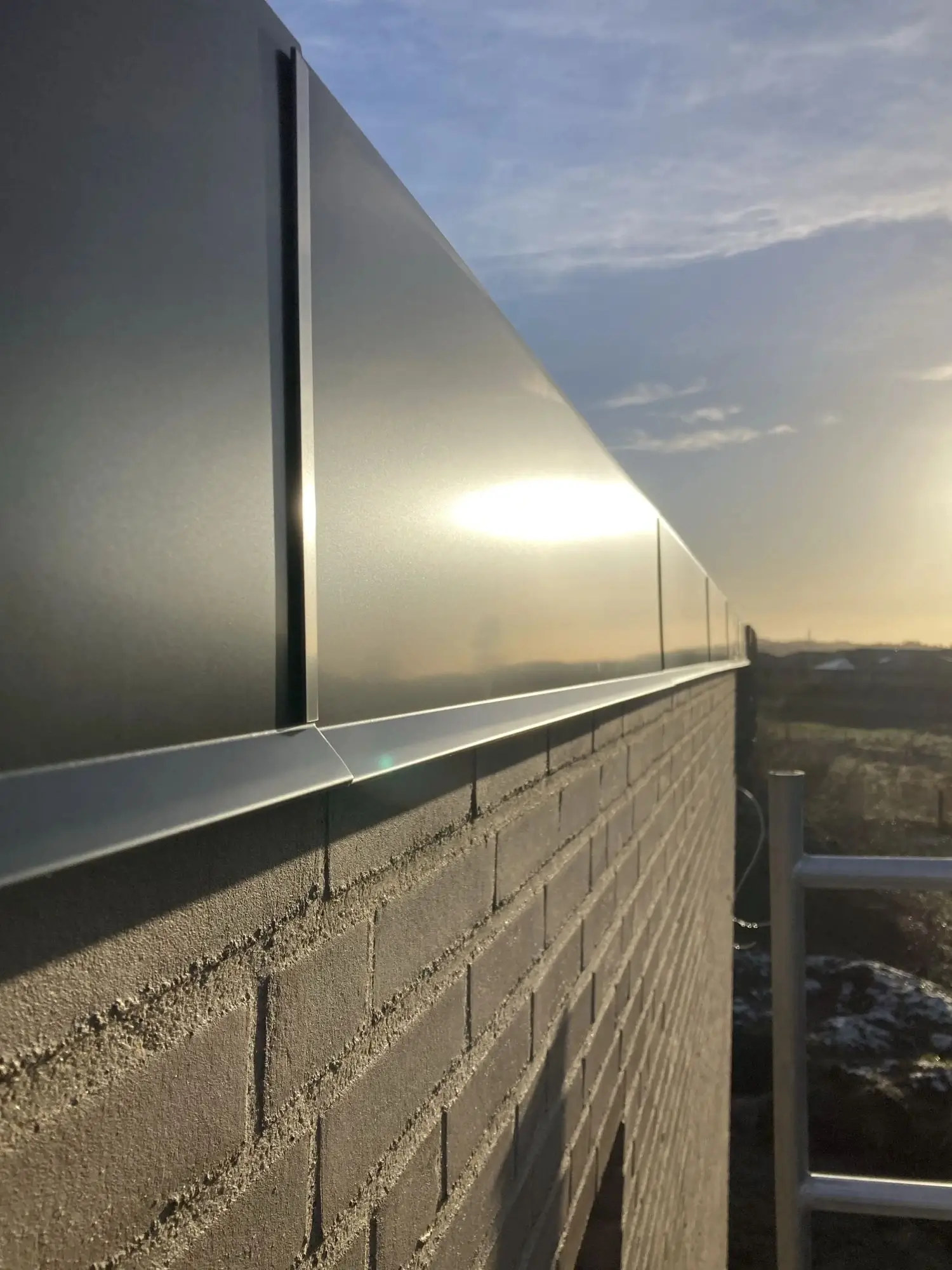 A close-up view of a brick wall topped with reflective metal panels under a partly cloudy sky at sunrise or sunset.
