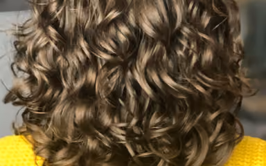 Back view of medium-length, thick brown hair styled in loose curls over a yellow sweater.