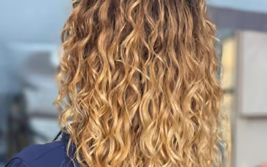 Back view of person with long, curly, highlighted blonde hair wearing a navy top indoors.