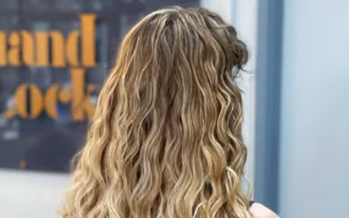 Back view of long wavy blonde hair in a salon with a blurred salon sign in the background.