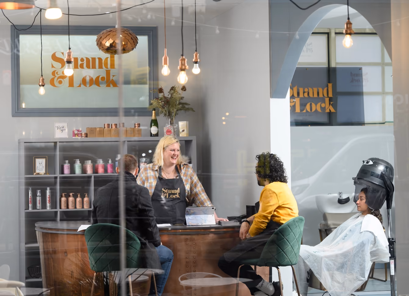 Salon receptionist smiles at two seated clients while a woman dries her hair under a hooded dryer.