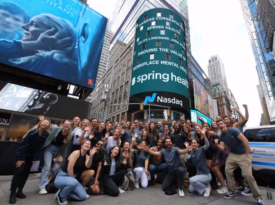A gathering of Spring Health employees in downtown Manhattan by a NASDAQ display on a building. The display reads "NASDAQ congratulates Spring Health on its landmark JAMA study proving the value of workplace mental health"