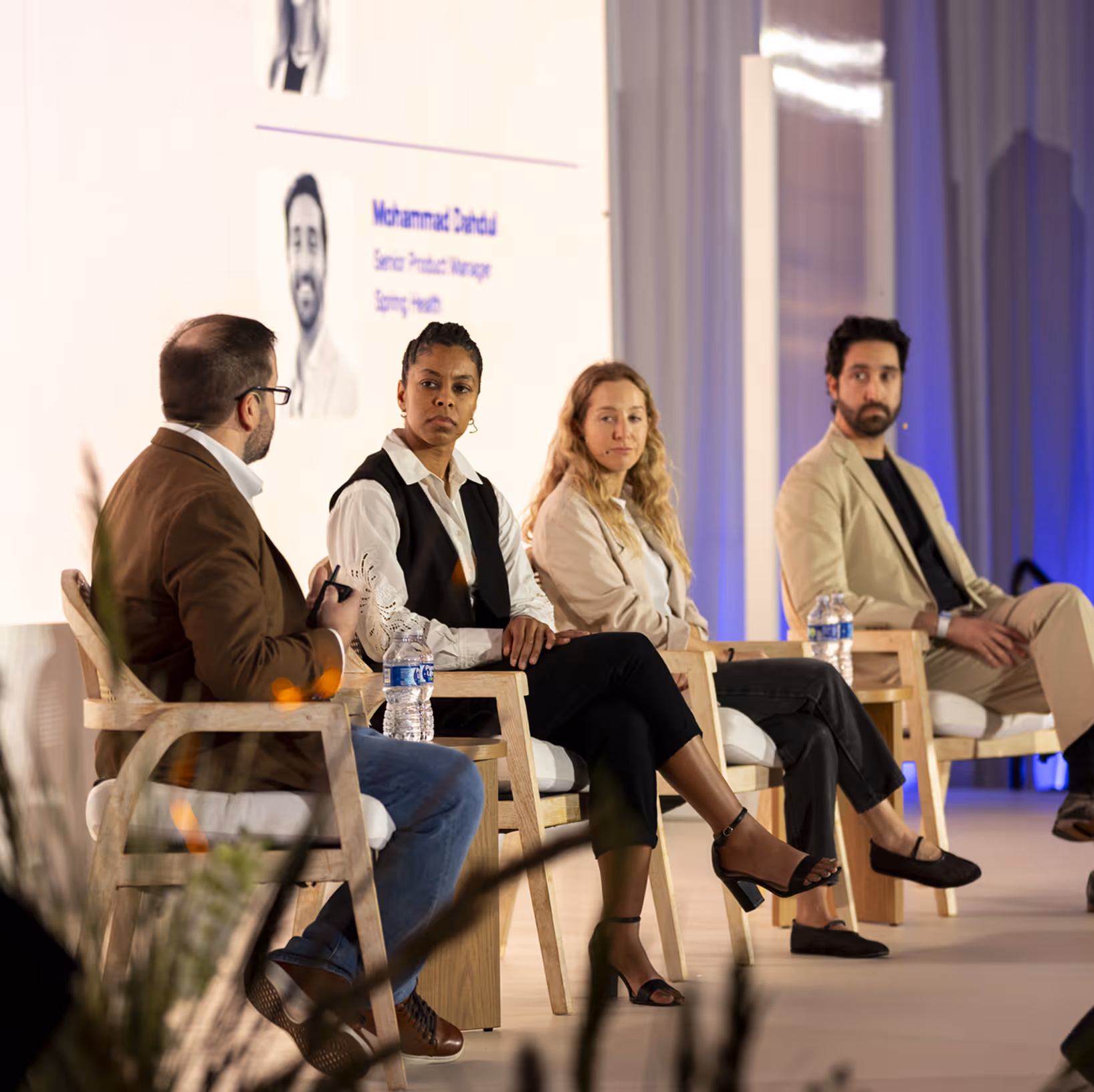 Four people seated in wooden chairs on a stage, engaged in a panel discussion at a conference.