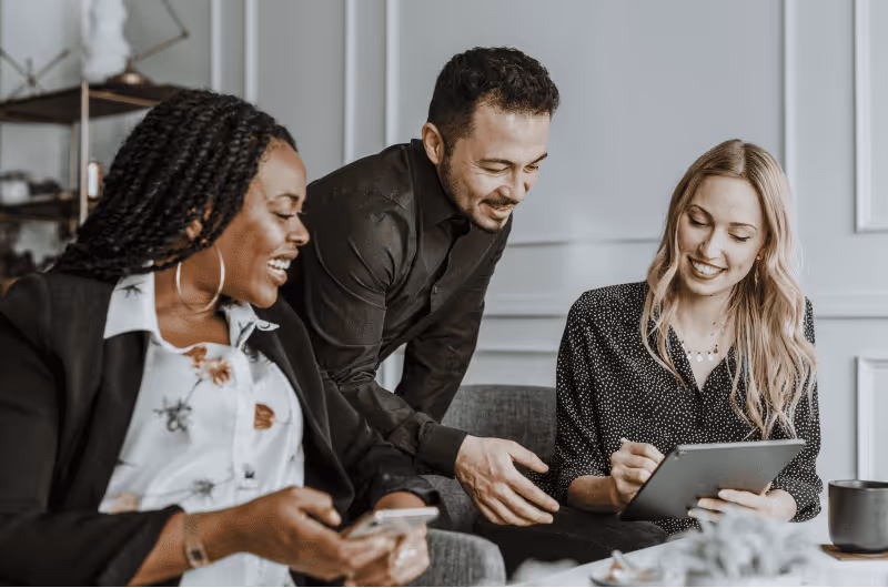 Three colleagues smiling and collaborating with a tablet and smartphone in an office setting.