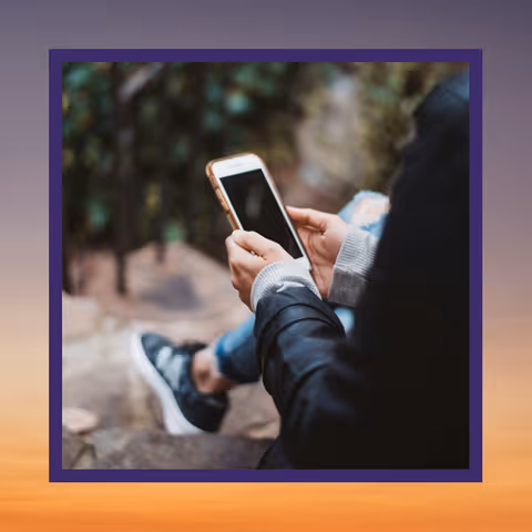 Person sitting outdoors holding and looking at a smartphone with a blurred natural background.