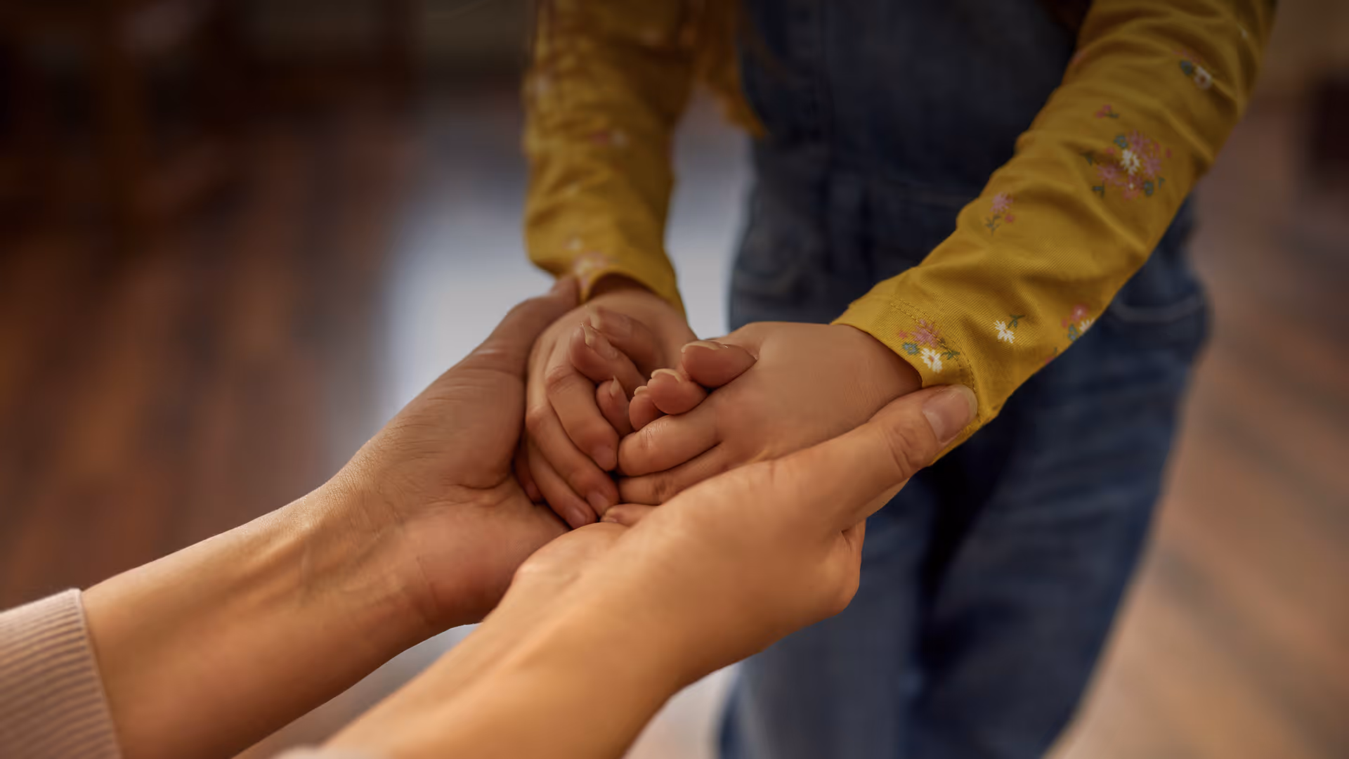Adult hands gently holding a child's clasped hands, child wears a yellow floral shirt and denim overalls.