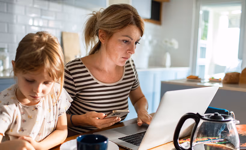 Woman focused on laptop while holding phone with child sitting beside her at kitchen table.