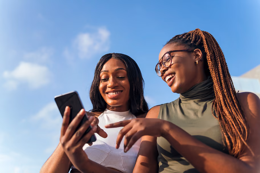 Two smiling young women outdoors looking at a smartphone against a blue sky.