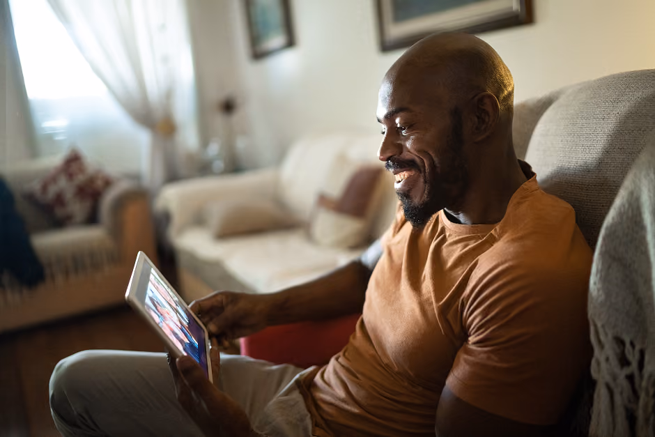 Smiling man sitting on a couch video chatting on a tablet in a cozy living room.