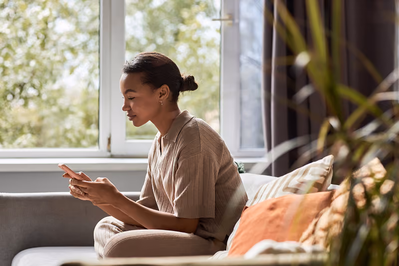 Woman sitting on a couch looking at her phone in a sunlit room with plants and cushions.