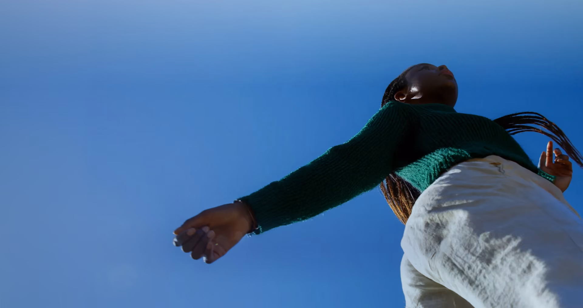 Low-angle view of a person in a green sweater with long braids against a clear blue sky.