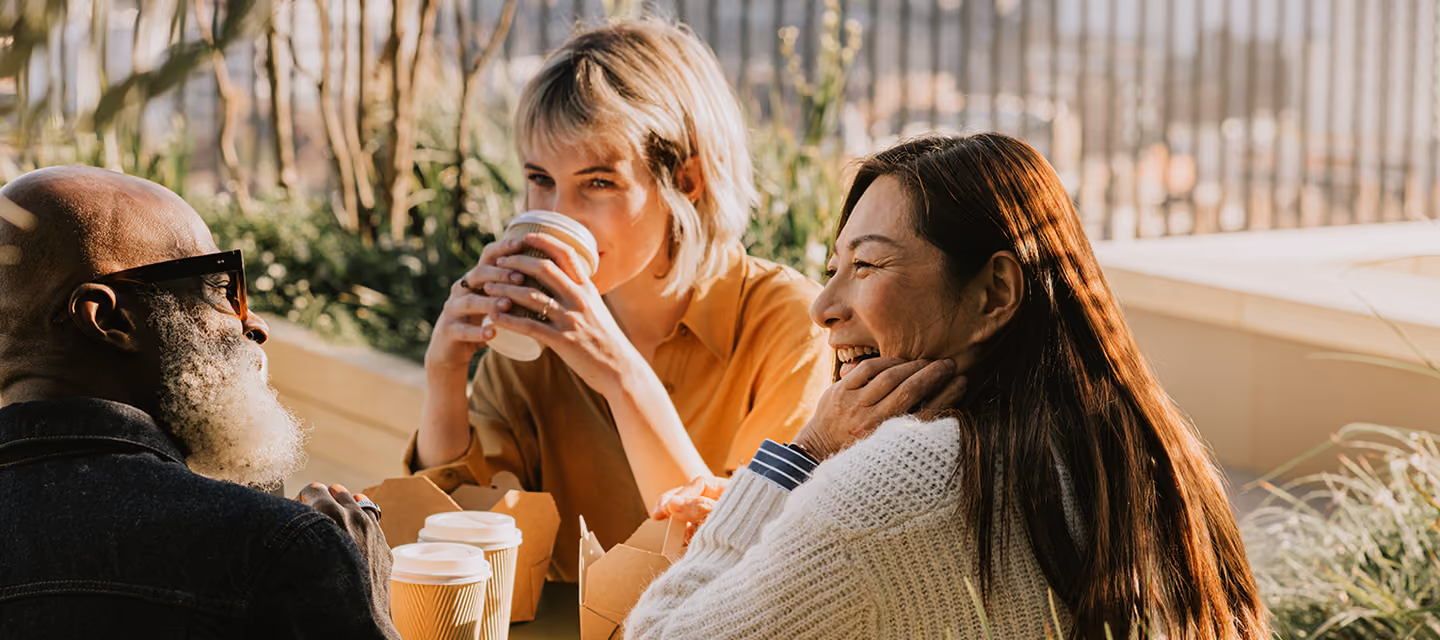 Three people enjoying coffee together outdoors with takeaway cups and food containers, smiling and engaging in conversation on a sunny day.