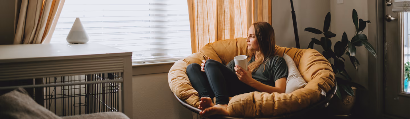 A woman with long hair sits curled up in a round cushioned chair, holding a white mug and looking out a window with blinds and beige curtains in a cozy room.