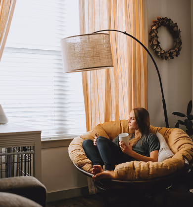 Woman sitting comfortably in a large round cushioned chair near a window with beige curtains, holding a white mug and looking thoughtfully outside while a tall floor lamp arches over her.
